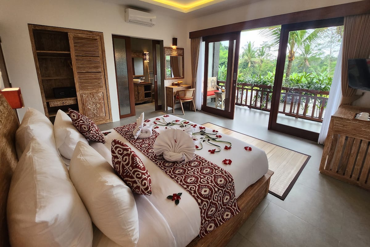 A hotel room with a bed, a desk, a chair, a TV, and a balcony with a view of greenery.