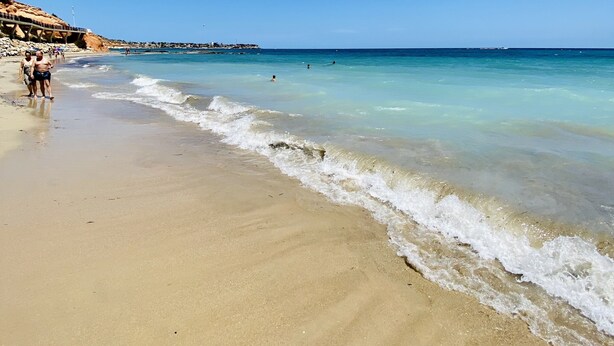 Una playa con agua azul cristalina, orilla de arena y algunas personas caminando.