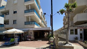 Un edificio de varios pisos con balcones, un área de estar techada con sillas y un pequeño árbol en una maceta.