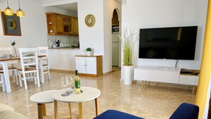 Una sala de estar moderna con un sofá azul, un comedor con sillas blancas, una cocina con armarios de madera y un televisor de pantalla plana montado en la pared.