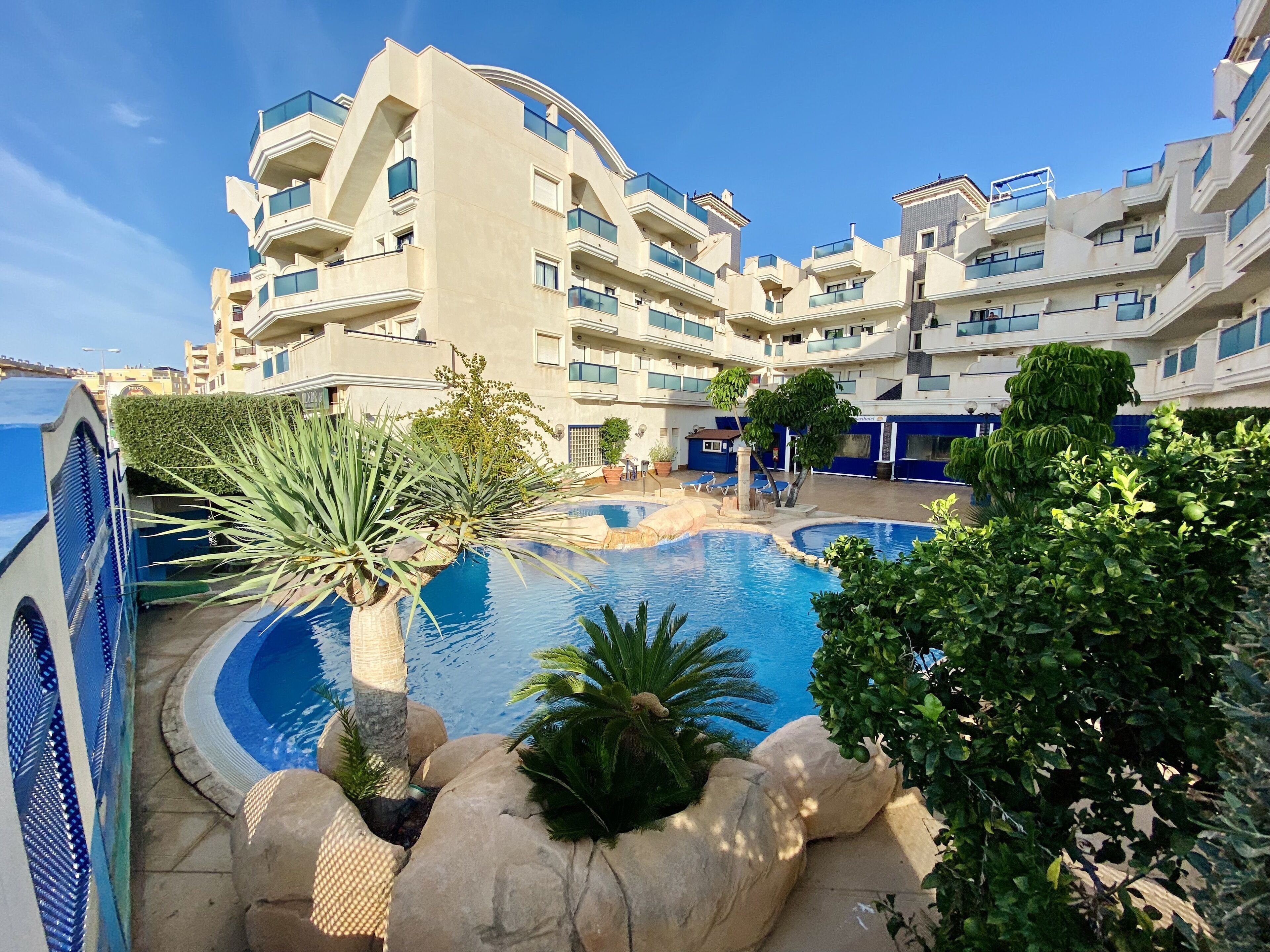 Un hotel moderno con zona de piscina rodeada de vegetación y un cielo azul despejado.