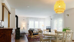 A spacious living room with a wooden dresser, a sofa, a dining table with chairs, and a large window with curtains. - Sunstreet by Interhome (Balatonoszod)