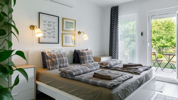 A modern bedroom with a bed, bedside tables, wall-mounted lamps, framed artwork, and a view of the outdoors. - Sunstreet by Interhome (Balatonoszod)