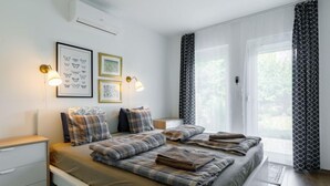 A bedroom with a large bed, a bedside table, a nightstand, and a window with curtains. - Sunstreet by Interhome (Balatonoszod)
