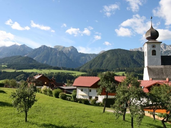 A rural village with a church and houses with red roofs, surrounded by green fields and mountains.