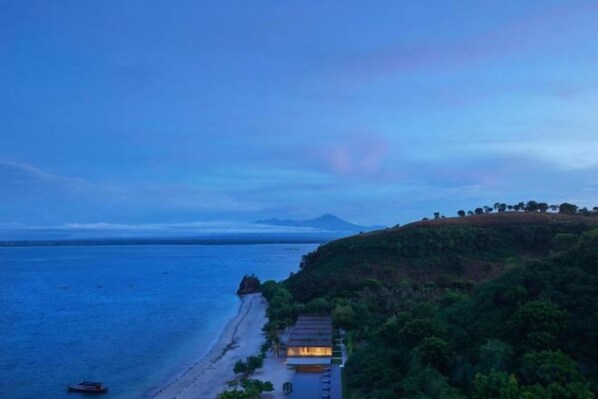 A coastal view with a beach, a small building, and a hill with trees.