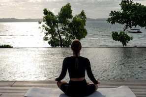 Una persona medita su un tappetino da yoga vicino a un lago, con alberi e una barca sullo sfondo.