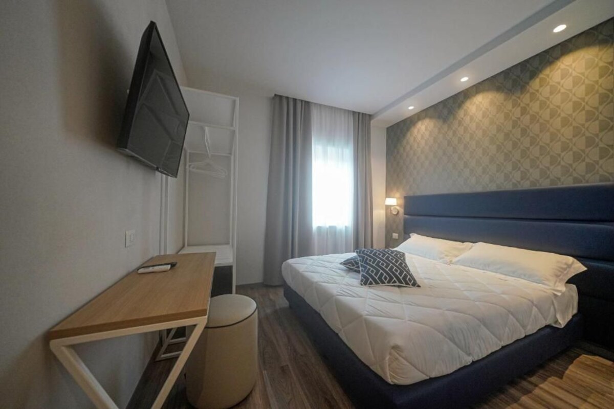 A hotel room with a bed, a desk, a chair, a TV mounted on the wall, and a window with curtains.