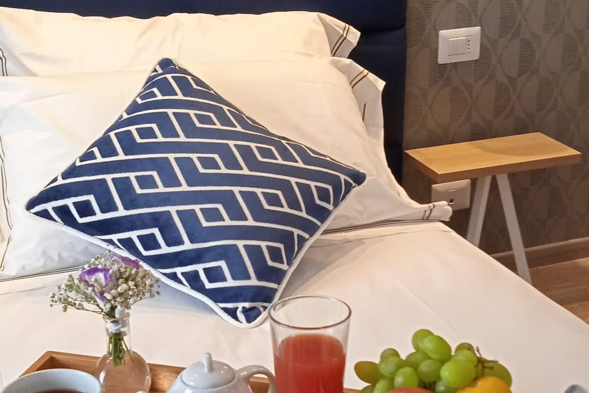 Un plateau de petit-déjeuner bien présenté, avec des croissants, des fruits, des céréales et des boissons, posé sur un lit doté d’une tête de lit bleue et d’un papier peint à motifs.