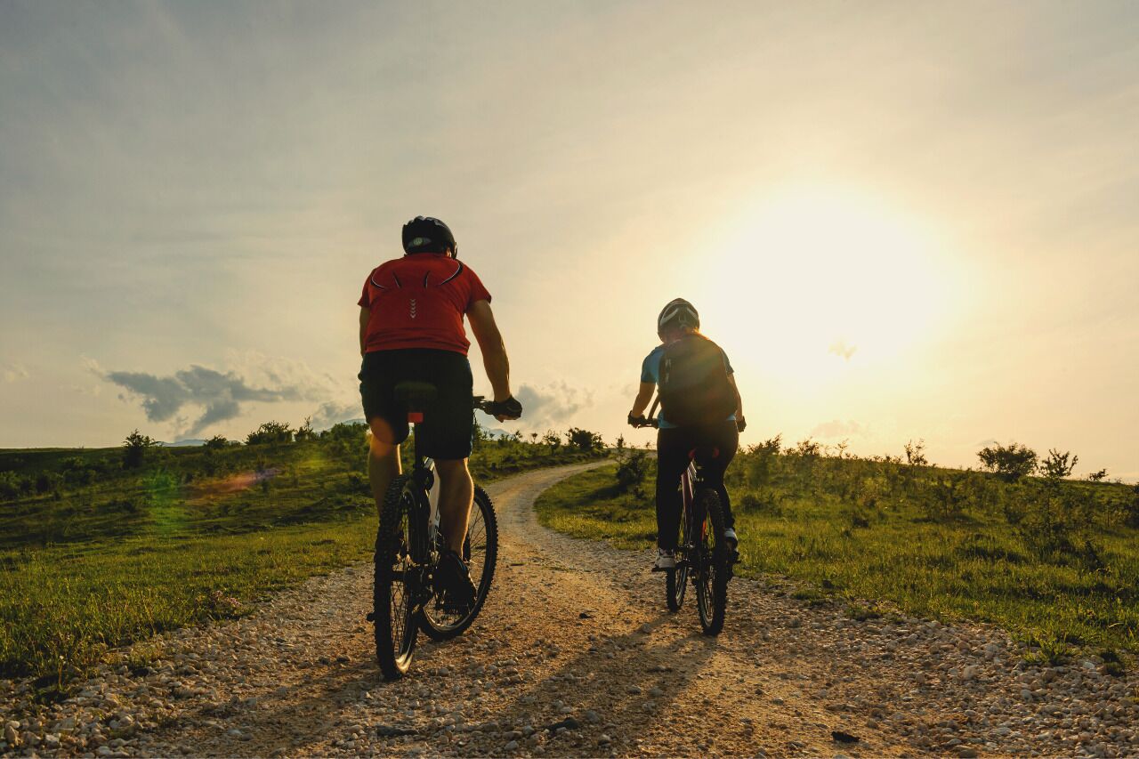 Dos personas montando en bicicleta por un sendero de grava con el atardecer de fondo.