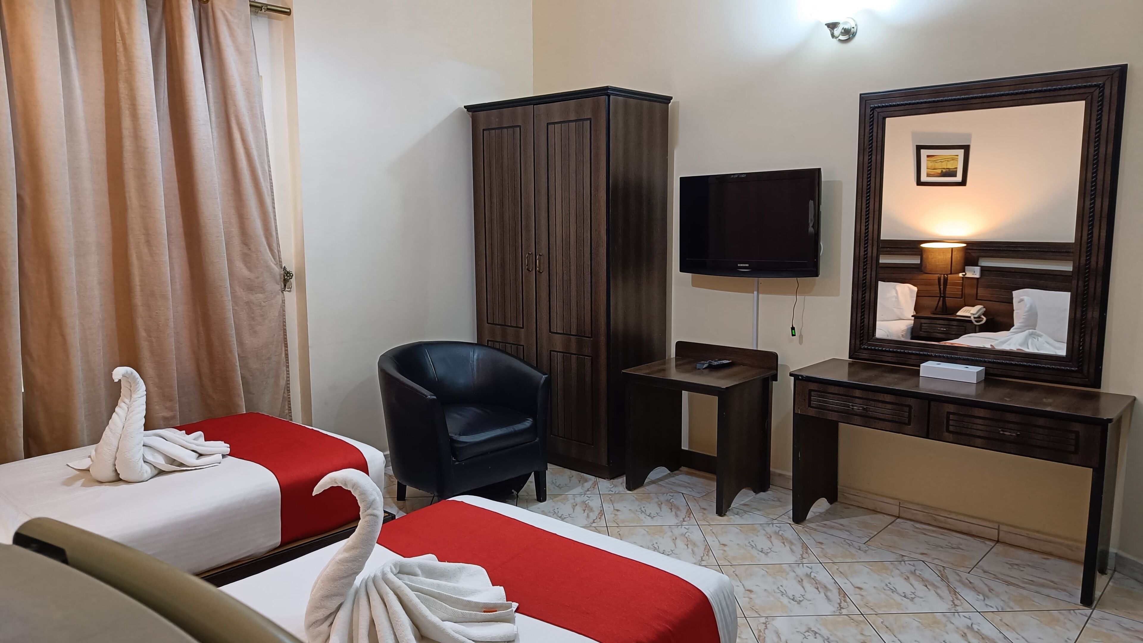 A hotel room with two beds, a wardrobe, a TV, a bedside table, a mirror, and a lamp.