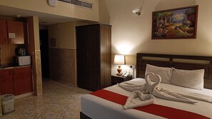 A hotel room with a bed, a nightstand with a lamp, a painting on the wall, and a kitchenette area.