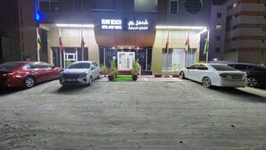 A hotel entrance with a sign reading "Ruwi Beach Hotel Apartments" and multiple flags. There are three cars parked in front. - RUWI BEACH HOTEL APARTMENTS (Sharjah)