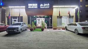Een hotelentree met een bord waarop 'RUWI BEACH HOTEL APARTMENTS' staat en verschillende vlaggen. Er staan twee auto's geparkeerd voor de ingang.