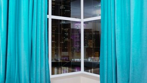 A corner room with teal curtains, a black chair, a wooden table, and a city view through a large window. - RUWI BEACH HOTEL APARTMENTS (Sharjah)