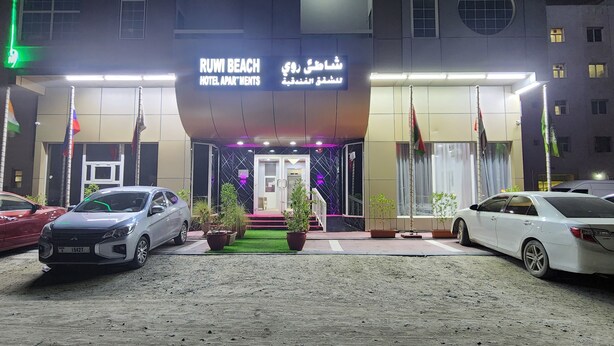 A hotel entrance with a sign reading "RUWI BEACH HOTEL APARTMENTS" and multiple flags. There are cars parked in front and potted plants along the walkway. - RUWI BEACH HOTEL APARTMENTS (Sharjah)