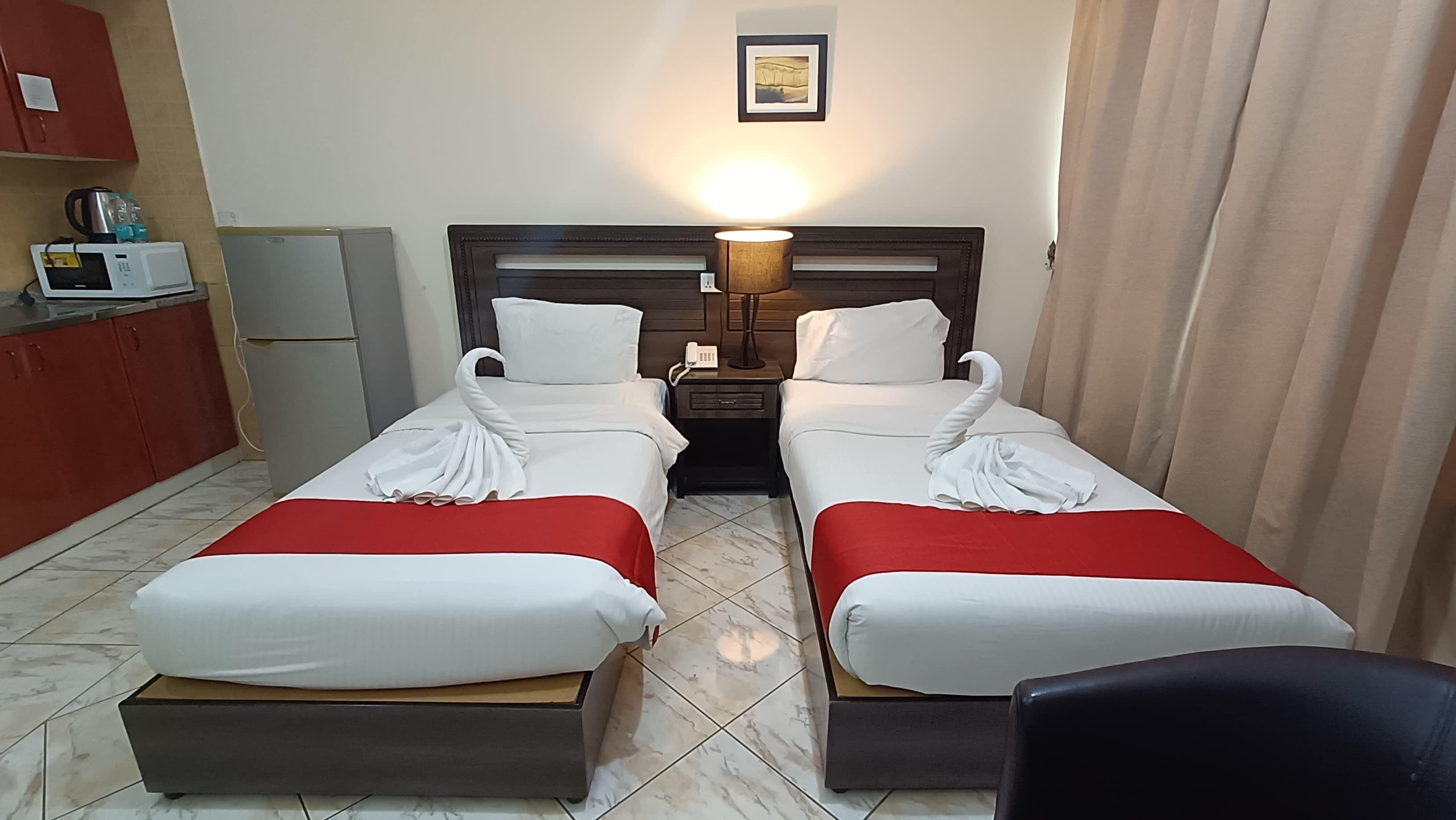 Een hotelkamer met twee bedden, een klein kitchenette, een bedlampje en een ingelijst schilderij aan de muur.