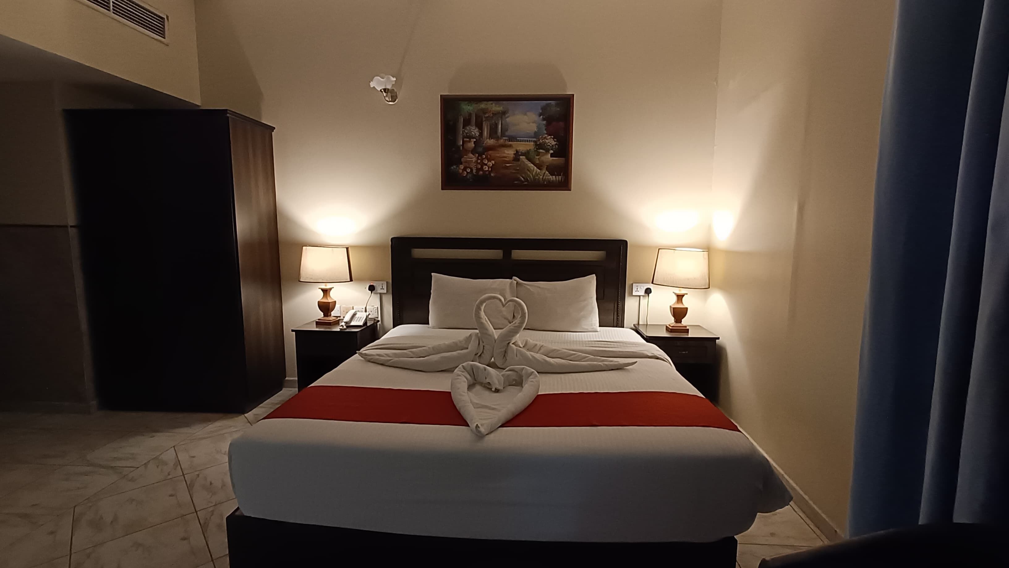 Habitación de hotel con una cama bien hecha, dos lámparas de noche, un cuadro en la pared y un ventanal con cortinas.