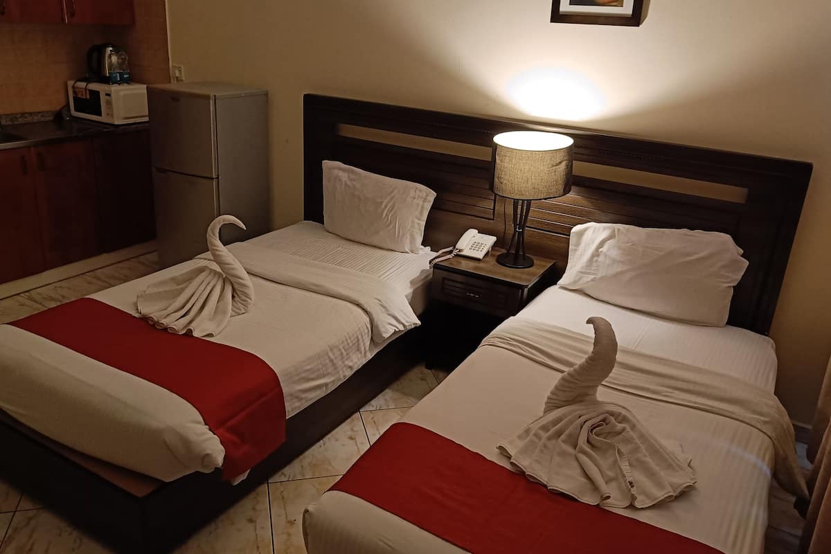 A hotel room with two beds, each with a towel shaped like a swan, a bedside table with a lamp, and a small kitchen area with a microwave and kettle.