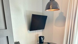 Une chambre d’hôtel moderne avec un fauteuil gris, une table ronde sur laquelle se trouve une tasse à café noire, une télécommande et un téléviseur fixé au mur.