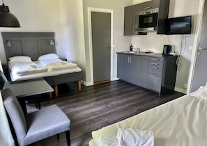 Habitación de hotel pequeña con cama, escritorio, silla, kitchenette y televisor montado en la pared.