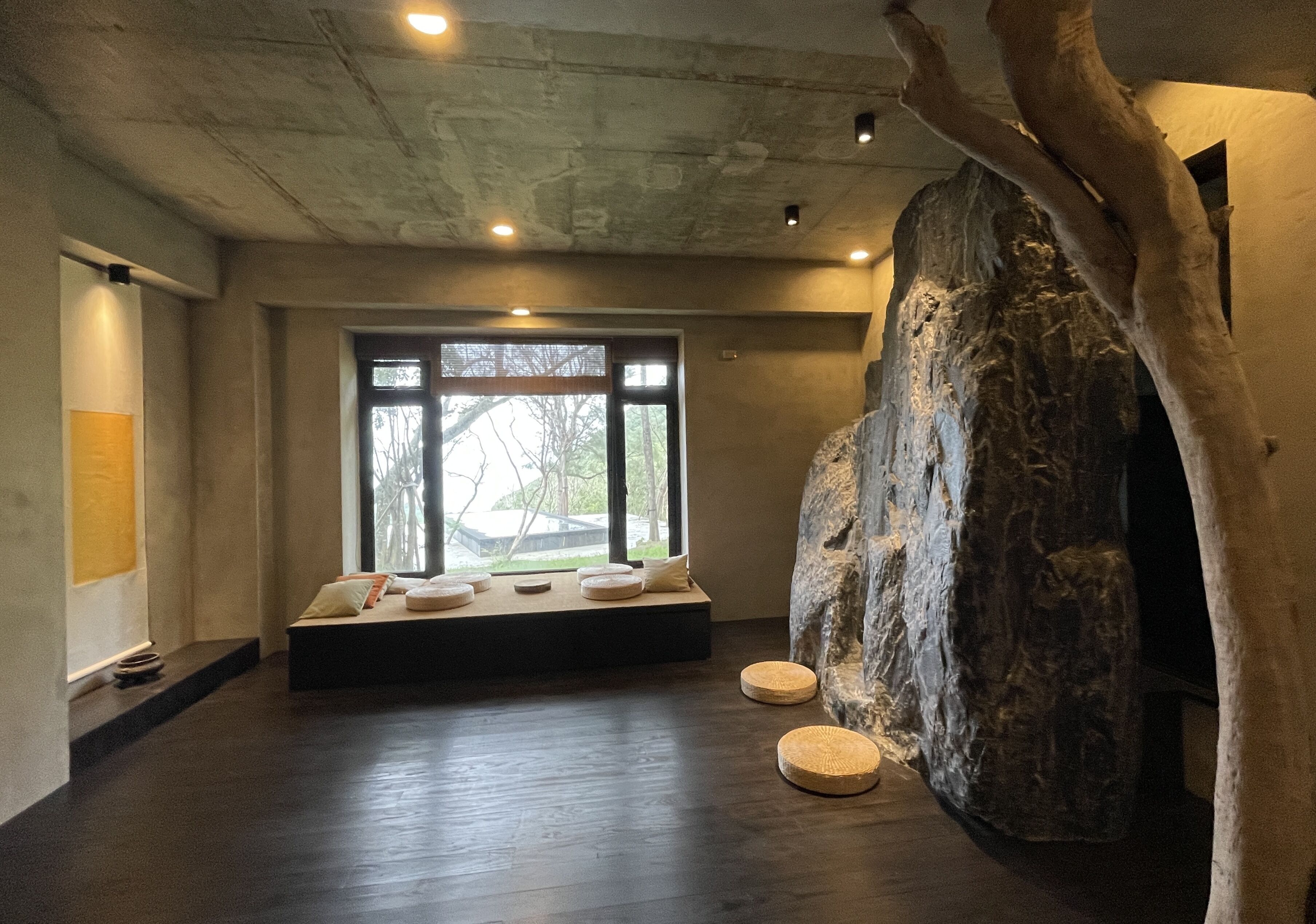 A spacious room with a large window, a wooden tree sculpture, and a seating area with cushions.