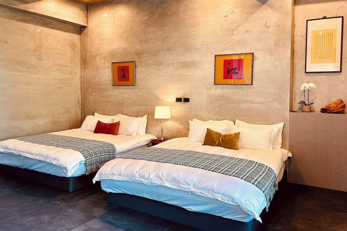 A hotel room with two beds, a small table, and a wall-mounted lamp.