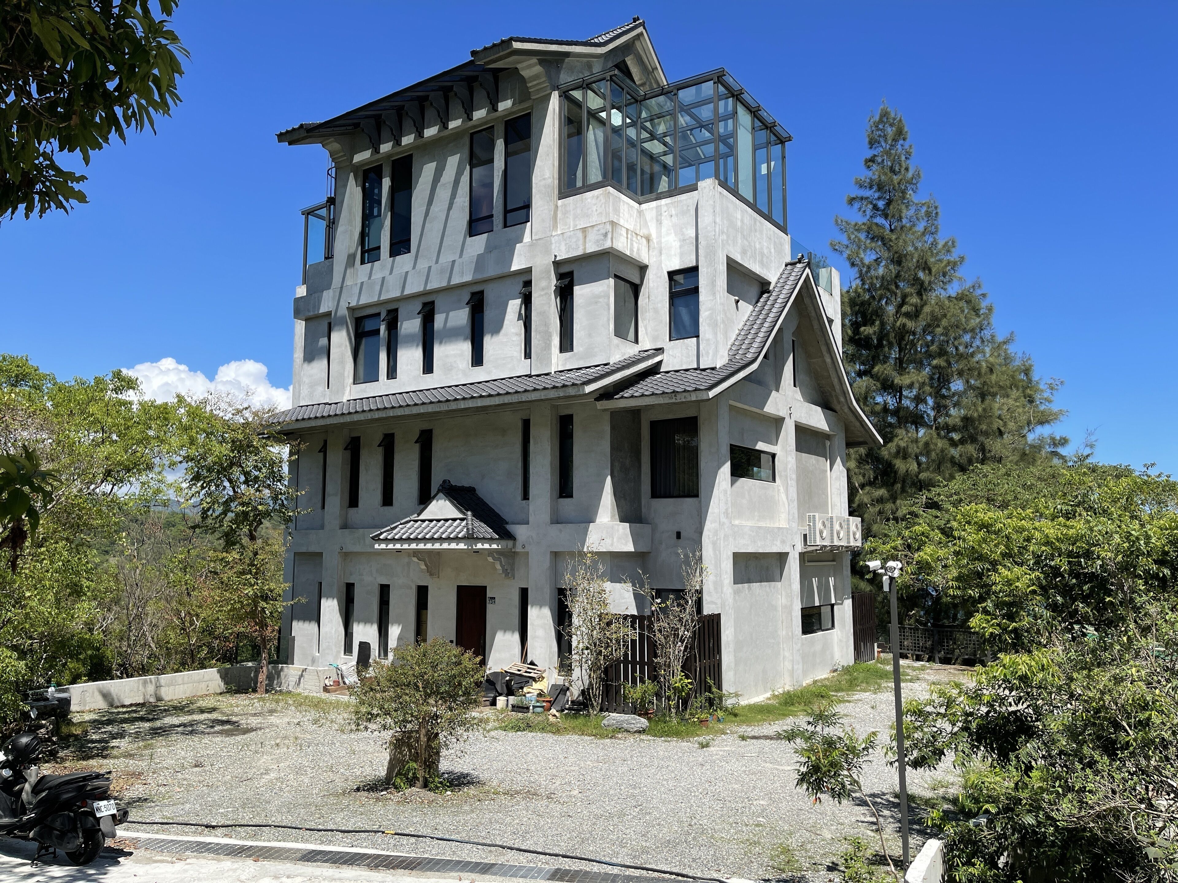 A modern building with a glass upper floor and a traditional lower section, surrounded by trees and a gravel driveway.