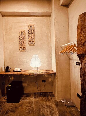 Een kleine, zwak verlichte kamer met een houten bureau, een lamp en een kapstok. Twee decoratieve panelen sieren de muur en er staan een paar schoenen op de vloer.