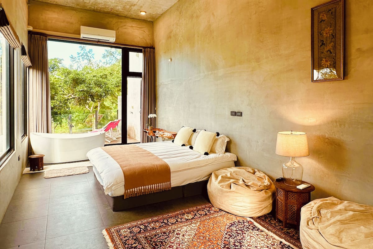 A bedroom with a large bed, a sitting area with a chair, a small table, a lamp, and a view of the outdoors.