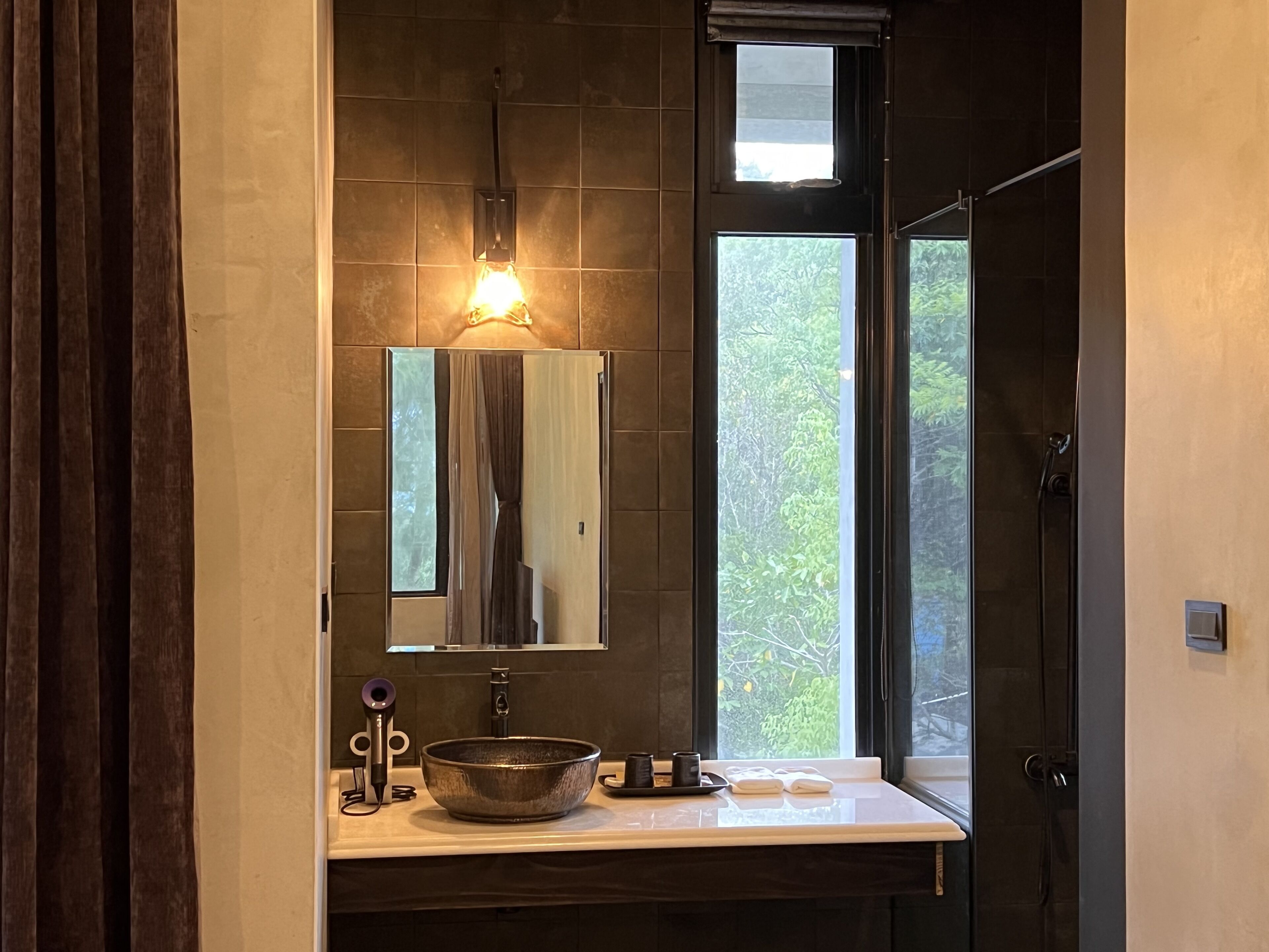 Un baño moderno con un espejo grande, un lavabo tipo recipiente y una ventana con vista a follaje.