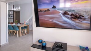 Una sala de estar moderna con un televisor de pantalla plana grande, una zona de comedor con sillas azules y un aparador con objetos decorativos.