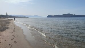 Una playa con una persona caminando por la orilla, un velero a lo lejos y una montaña al fondo.