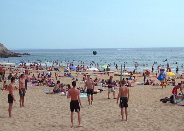 Una playa concurrida donde gente toma el sol y juega al voleibol.