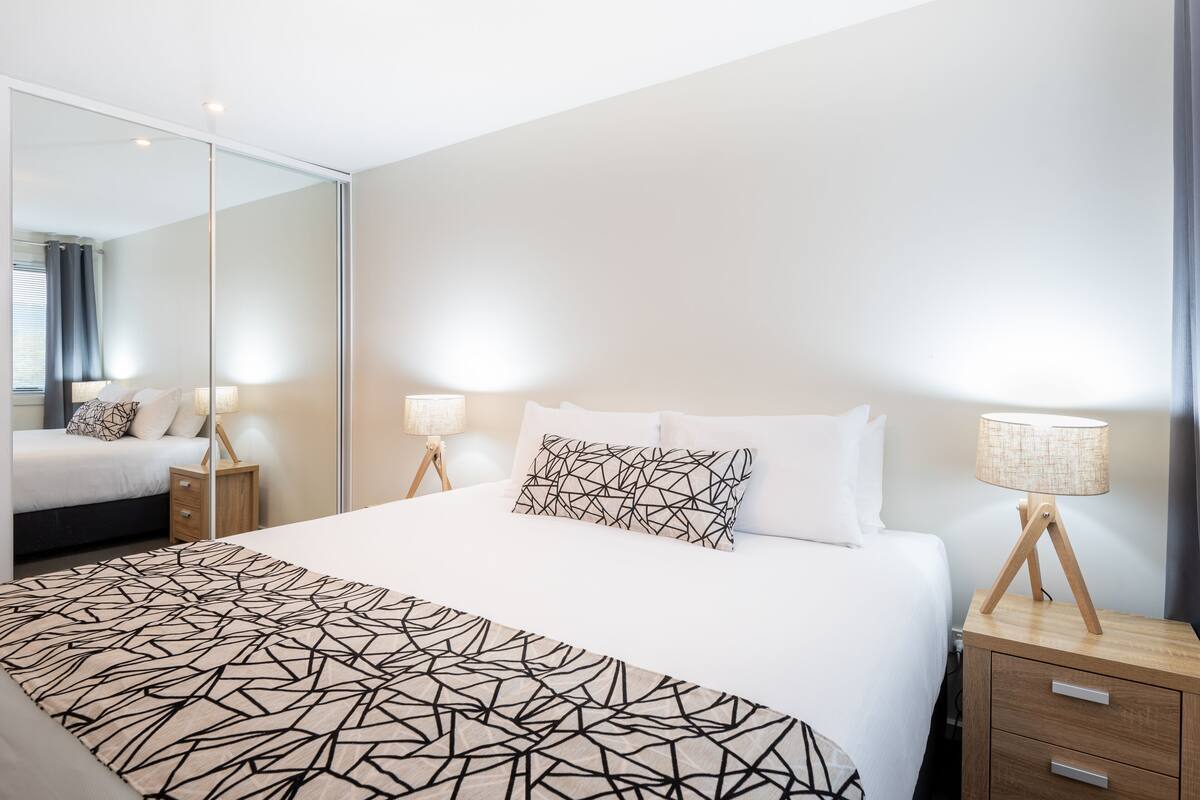 A modern hotel room with a large bed, bedside tables, a mirror, and a lamp.
