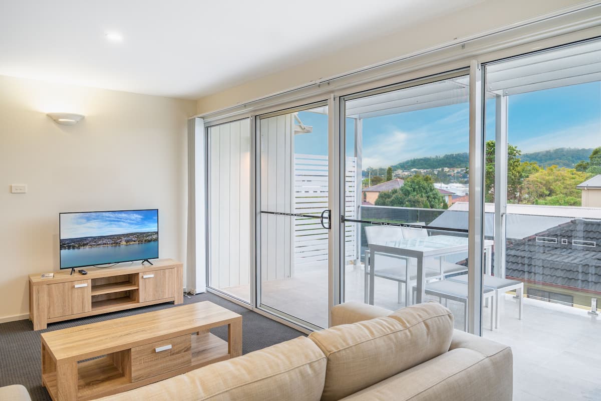 A modern living room with a flat-screen TV, wooden entertainment unit, sofa, and a glass door leading to an outdoor balcony with a view of the city and hills.