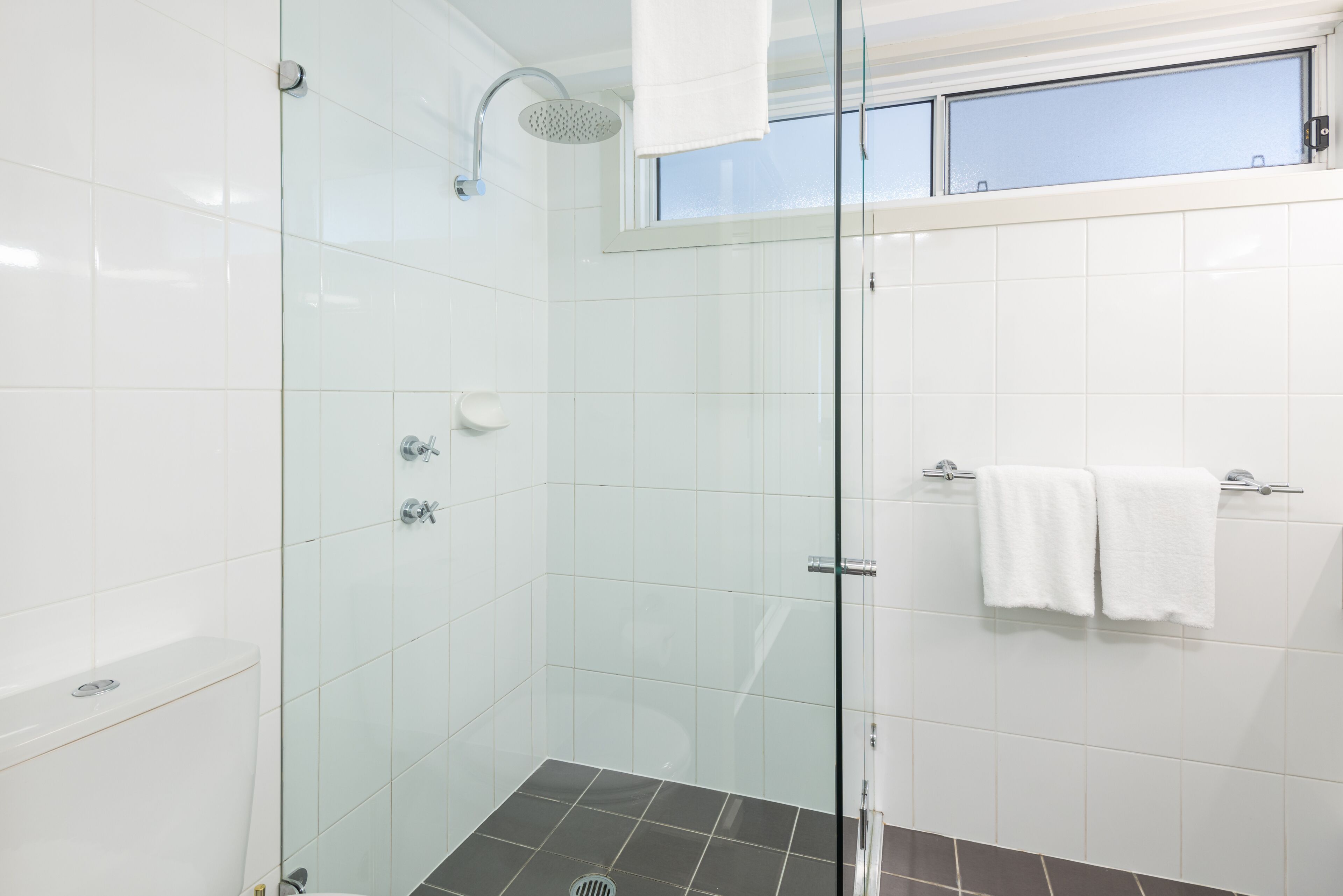 A modern bathroom with a glass shower enclosure, white tiles, and a window above the sink.