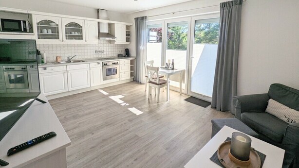 Cozinha moderna com armários brancos, área de jantar com mesa e cadeiras, e sofá cinza.