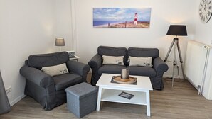 Uma sala de estar com dois sofás cinzas, uma mesa de centro branca, um abajur, um quadro de um farol e um relógio na parede.