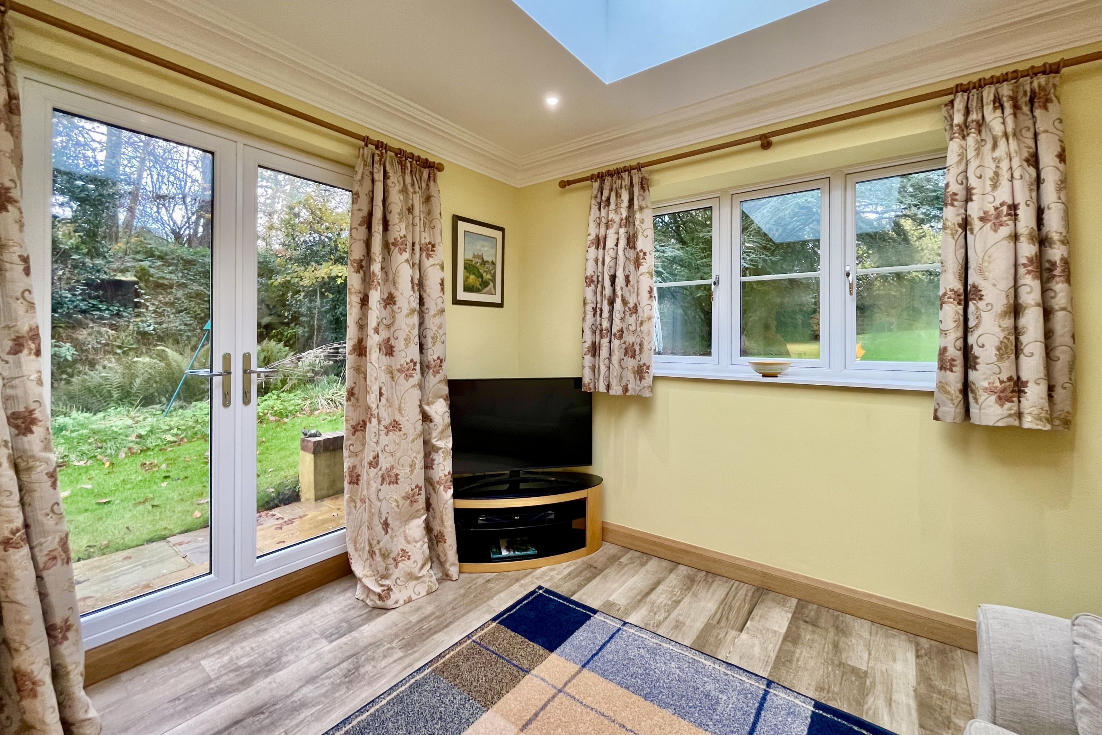 A room with a TV on a stand, a large window with curtains, and a view of a garden.