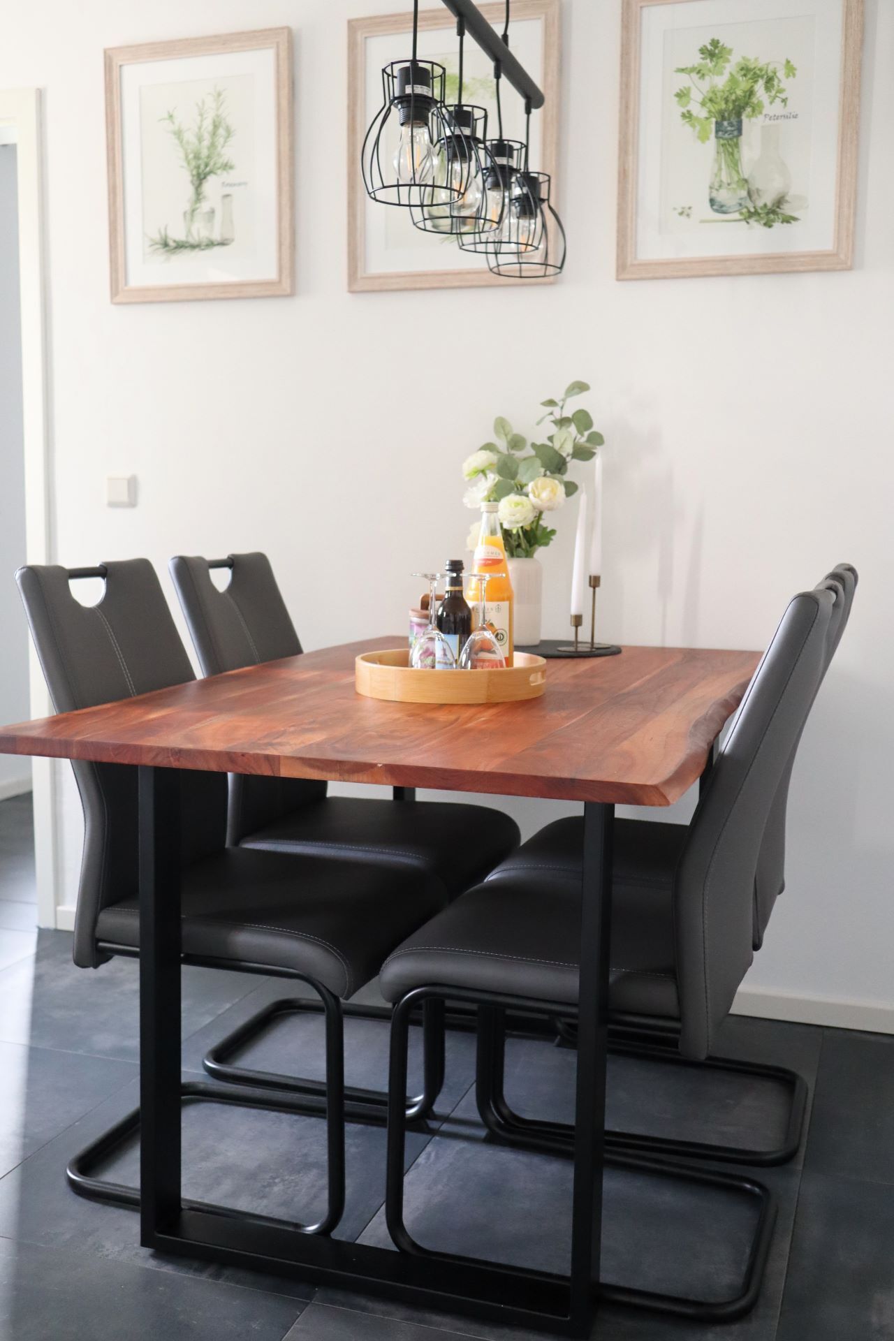 Une table à manger avec un plateau en bois et un pied en métal noir, entourée de chaises en cuir noir. Au-dessus de la table, on peut voir trois illustrations botaniques encadrées accrochées au mur.