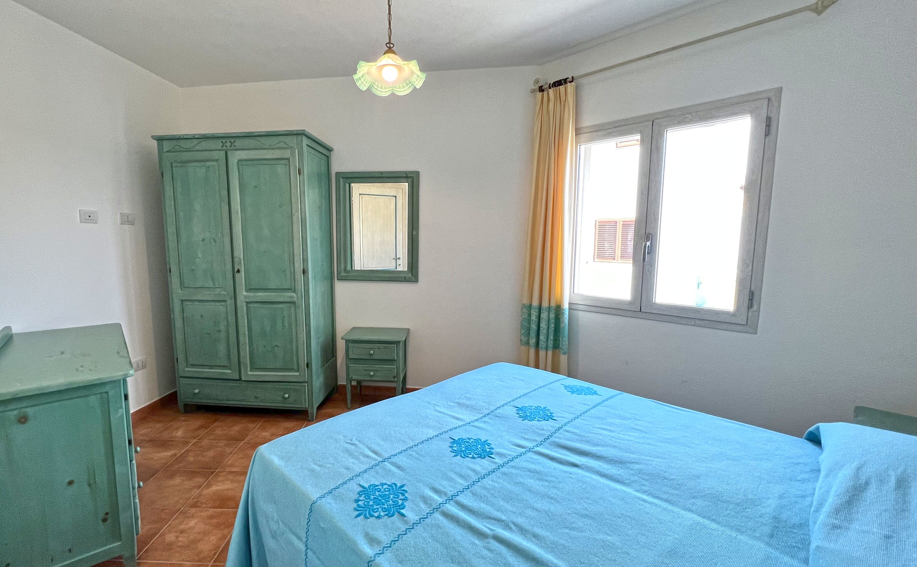 Un dormitorio con cama, armario, mesita de noche, espejo y ventana con cortinas.