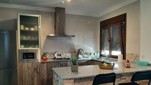 Una cocina moderna con microondas integrado, cocina a gas y refrigerador de acero inoxidable. Hay dos sillas negras en la isla y una ventana con persianas.