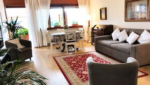 Una sala de estar con un sofá gris, un tapete rojo y blanco, una mesa de comedor con sillas y un piano.