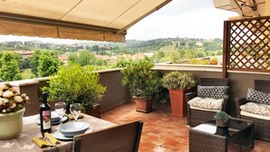 Una terraza con mesa dispuesta para dos, plantas en macetas, mobiliario de mimbre y vistas a la ciudad.