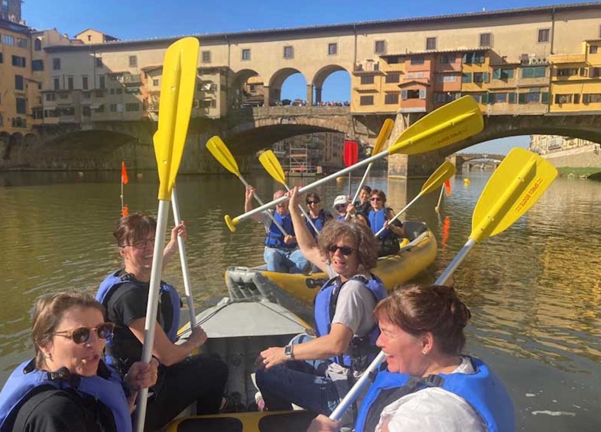 A group of people on a boat with yellow paddles, wearing life jackets, and a bridge in the background.