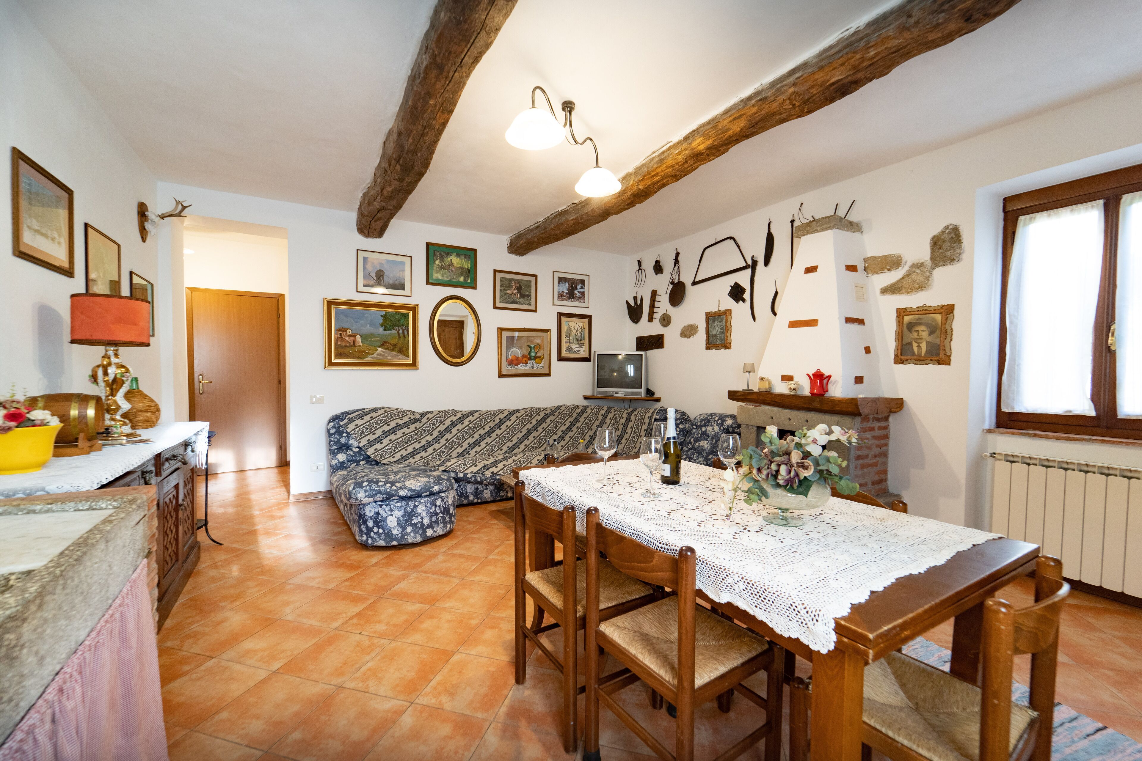 Un salon chaleureux avec une table à manger en bois, un canapé et un téléviseur. Des tableaux encadrés ornent les murs et une lampe est posée sur la table.