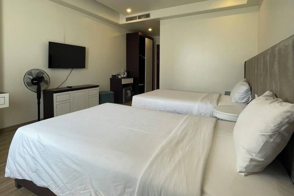 A hotel room with two beds, a flat-screen TV mounted on the wall, a desk with a chair, and a standing fan.