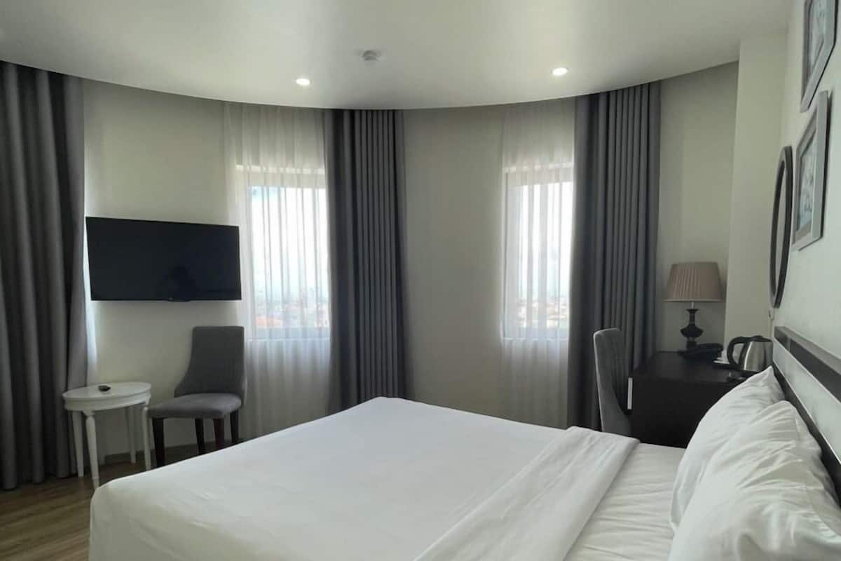 A hotel room with a large bed, a flat-screen TV mounted on the wall, a small desk with a chair, and a lamp on a nightstand.