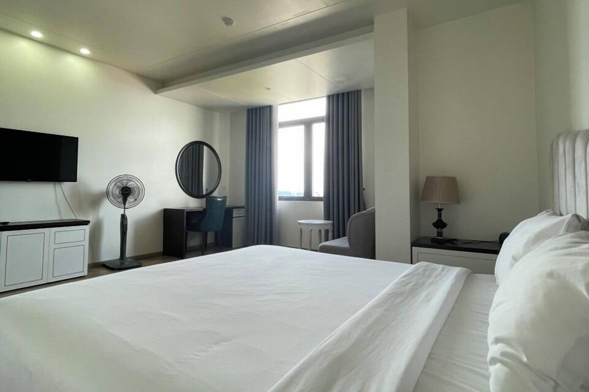 A modern hotel room with a large bed, a desk, a chair, a round mirror, a lamp, and a TV mounted on the wall.
